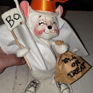 Puede incluir: Un rat&oacute;n de fieltro blanco vestido de fantasma para Halloween. El rat&oacute;n lleva un sombrero de copa blanco con una cinta naranja, un disfraz de fantasma blanco y sostiene una bolsa de papel marr&oacute;n que dice "Trick or Treat". El rat&oacute;n tambi&eacute;n sostiene un cartel blanco que dice "Boo".