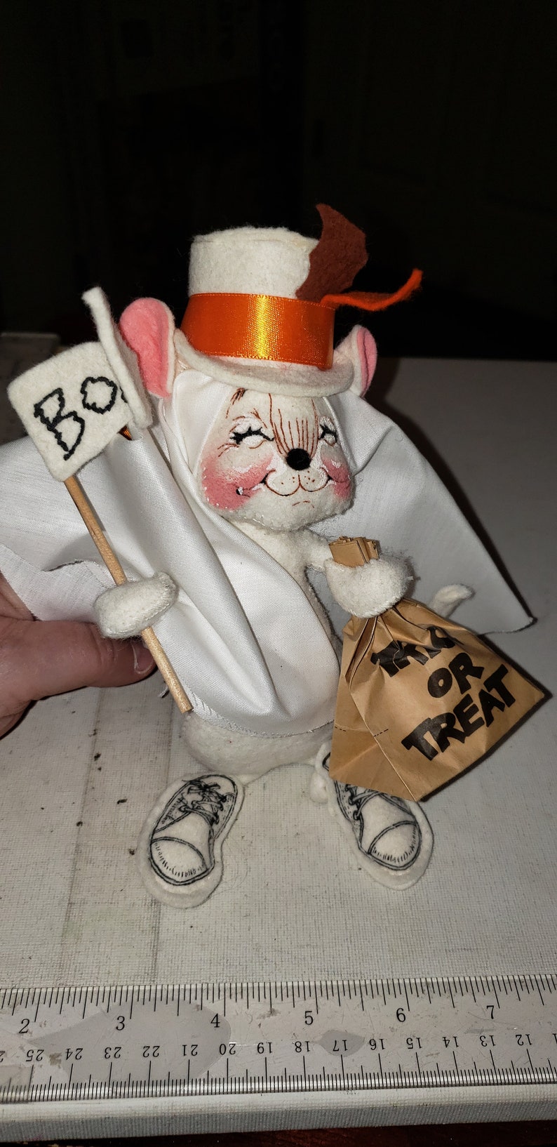 Puede incluir: Un rat&oacute;n de fieltro blanco vestido de fantasma para Halloween. El rat&oacute;n lleva un sombrero de copa blanco con una cinta naranja, un disfraz de fantasma blanco y sostiene una bolsa de papel marr&oacute;n que dice "Trick or Treat". El rat&oacute;n tambi&eacute;n sostiene un cartel blanco que dice "Boo".
