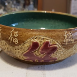 May include: A brown ceramic bowl with a green glaze inside. The bowl has a raised design of Chinese characters and a red floral pattern.