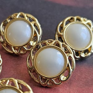 May include: Close-up of several gold-coloured buttons with a white, round centre. The buttons have a decorative, openwork design around the centre. These buttons are likely for clothing or craft projects.