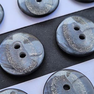 May include: A collection of round, dark gray buttons featuring a smooth, glossy finish. Each button has a central band of silver glitter texture and two holes for attachment. The buttons are arranged in rows on a white and black surface.