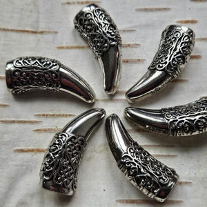 Puede incluir: Seis colgantes plateados en forma de garra dispuestos en un patrón circular. Cada colgante presenta un diseño ornamentado y grabado en la parte superior y una punta lisa y curvada. Los colgantes se exhiben sobre una superficie clara y texturizada.