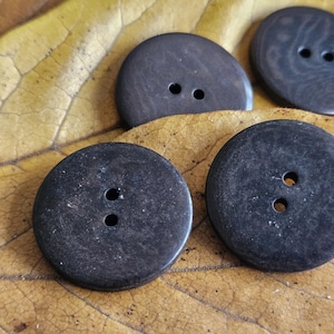 Pode incluir: Close-up de vários botões redondos castanhos escuros com dois furos cada, repousando sobre uma folha seca amarelo-castanha. Os botões têm uma superfície ligeiramente texturizada e um acabamento mate, sugerindo um material natural.