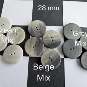 May include: A collection of round buttons in cream, beige, and grey mixes, each with two holes. The buttons are 28 mm in diameter. The image is on a black and white checkered background.