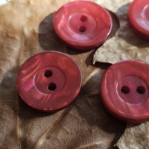 Può includere: Un set di bottoni rotondi a due fori in una tonalità rosso intenso e brillante. Ogni bottone presenta un centro liscio e leggermente concavo con una finitura lucida che riflette la luce, mettendo in risalto la forma circolare e i due fori.