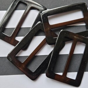 May include: Four dark brown rectangular belt buckles with a glossy finish. The buckles have a rectangular frame with a single bar across the middle. The background is black and white striped.