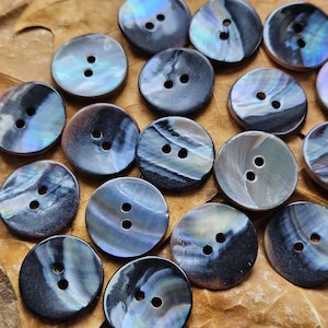 Puede incluir: Una colección de botones redondos e iridiscentes, cada uno con dos agujeros. Los botones tienen un patrón arremolinado y veteado en tonos de azul, gris y negro, con toques de morado y verde. Están dispersos sobre una superficie marrón y texturizada.