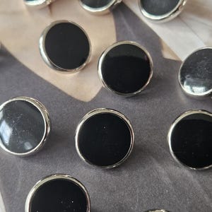 May include: A collection of round buttons with a black center and a silver-colored metal rim. The buttons are arranged on a patterned surface. These buttons could be used for clothing or craft projects.