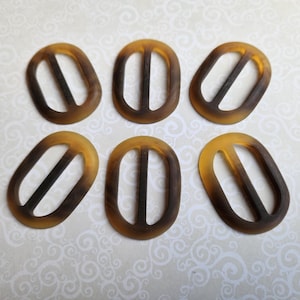 May include: Six oval belt buckles with a tortoiseshell design. Each buckle features a central bar and a gradient from light to dark brown. These accessories are suitable for various sewing and crafting projects.