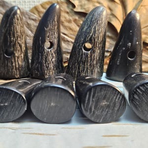 May include: A collection of dark, horn-shaped buttons with a natural, textured appearance. Each button has a hole for sewing. The buttons are arranged on a light surface, with some standing upright and others lying flat.