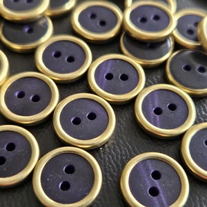 May include: A collection of round buttons featuring a deep purple center with a subtle sheen, each encased in a smooth, polished gold-toned metal rim. Every button has two centered holes for attachment, arranged in a uniform, circular design.