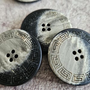 以下が含まれることがあります： 黒とグレーのマーブル模様が施された、中央に4つの穴がある3つの丸いボタン。各ボタンの周囲には、表面に彫り込まれたギリシャ雷文の縁取りが施されており、暗い色調と明るい色調の間に鮮明なコントラストを生み出しています。