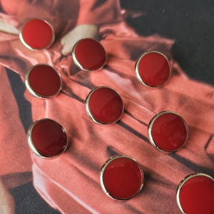 Könnte beinhalten: Zehn runde, rote Knöpfe mit silberfarbenen Rändern liegen auf einem strukturierten, rot-rosa Stoff. Die glänzenden Knöpfe sind über die Oberfläche verstreut. Diese Knöpfe sind ein klassisches Design, geeignet zum Nähen.