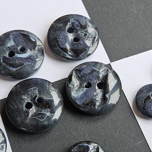 Può includere: Una collezione di bottoni grigio scuro con effetto marmorizzato. Ogni bottone ha due fori e un motivo a vortice di grigio chiaro e blu. I bottoni sono rotondi e di dimensioni variabili.