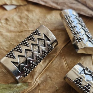 May include: Close-up of several silver-toned metal tubes with a decorative pattern. The pattern includes zig-zag lines and small, round, raised details. The tubes are arranged on a brown, textured surface, possibly a leaf.