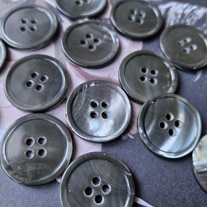 May include: A collection of gray, round buttons with four holes each. The buttons have a pearlescent sheen and are arranged on a patterned surface, suggesting a craft or sewing project. The buttons vary in size.