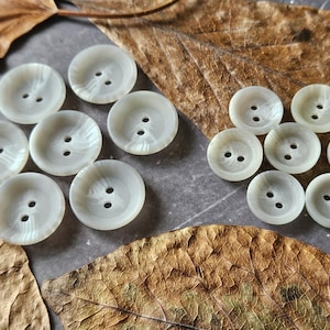 May include: A collection of off-white, round buttons with two holes each, arranged on a surface with dried leaves. The buttons have a subtle, marbled appearance, suggesting a pearlescent finish. Ideal for sewing and craft projects.