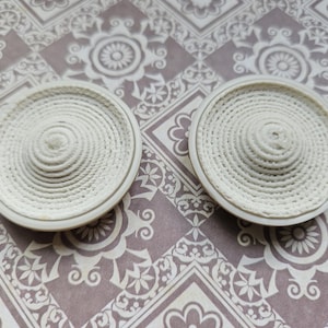 May include: Two off-white, circular coasters with a spiral rope design. The coasters have a raised centre and a rounded edge. They are placed on a patterned surface with floral and geometric elements.