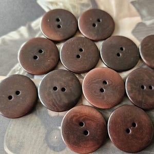 May include: A selection of dark brown, circular buttons, each with two holes. The buttons have a textured surface and a wood-grain effect. Ideal for sewing and craft projects. Each button is approximately 2.5 cm in diameter.