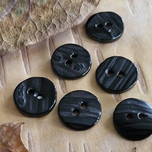 Può includere: Sei bottoni neri con effetto marmorizzato, ciascuno con due fori per cucire. I bottoni rotondi hanno una finitura lucida. Sono disposti su una superficie marrone chiaro strutturata. Ideali per progetti di cucito.