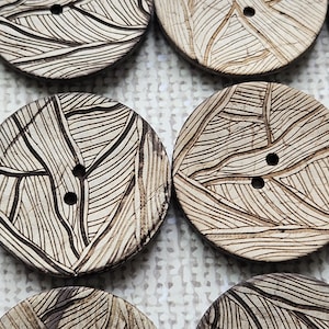 May include: Close-up of several round, wooden buttons with a leaf-like pattern etched into the surface. Each button has two holes for sewing. The buttons are light brown with darker brown etched lines.