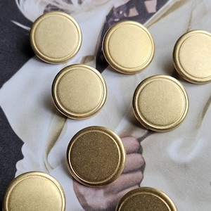 May include: A collection of eight round, brass-colored cabinet knobs. Each knob has a flat, circular face with a slightly raised, rounded edge. The knobs are arranged on a patterned background.