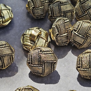 Puede incluir: Una colección de botones redondos de tono dorado con una textura tejida de mimbre. Cada botón tiene un acabado metálico en relieve con intrincados patrones de cordones entrecruzados y un vástago resistente en la parte posterior.