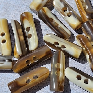 May include: A selection of oblong toggle buttons in shades of brown and cream. Each button has two holes for sewing. The buttons have a marbled appearance, with swirls of colour throughout. Suitable for various sewing projects.