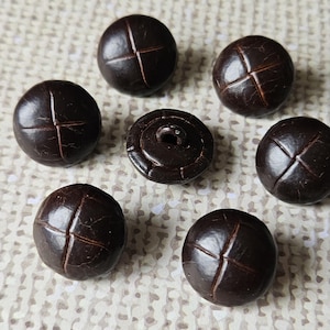 May include: Seven dark brown, round buttons with a cross-shaped design stitched into each. One button has a shank for sewing. The buttons are displayed on a light beige, textured surface. Each button is approximately 2 cm in diameter.