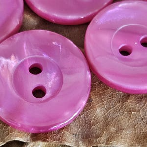 May include: A collection of round, bright pink buttons with a pearlescent, swirling finish. Each button features a recessed center with two circular holes for attachment. The surface of the buttons has a smooth, glossy texture with subtle light reflections.