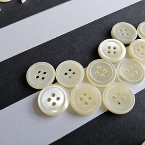 May include: A collection of off-white, pearlescent buttons with four holes each. The buttons are arranged on a black and white striped surface, with varying sizes and shapes. The buttons are made of a smooth, shiny material.