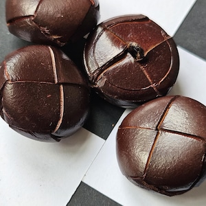 May include: Four dark brown leather buttons with a stitched design are arranged on a black and white checkered surface. The buttons have a rounded shape with a cross-like pattern created by the stitching.