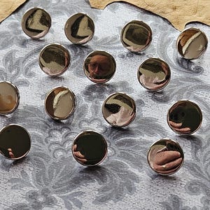 May include: A collection of twelve circular, metallic buttons with a highly reflective, polished silver finish. Each button features a smooth, convex surface that acts as a mirror, reflecting the surrounding environment and light sources clearly.