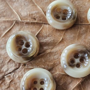 May include: Close-up of several round buttons with four holes each. The buttons have a marbled appearance with shades of cream and brown. They are arranged on a dried, brown leaf.