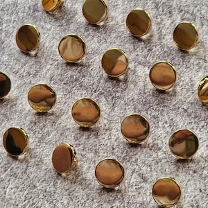 Könnte beinhalten: Eine Sammlung kleiner, runder, goldfarbener Metallknöpfe mit einer glatten, reflektierenden Oberfläche. Jeder Knopf hat ein einfaches, kreisförmiges Design mit poliertem Finish und ist verstreut angeordnet, um die einheitliche Form zu zeigen.