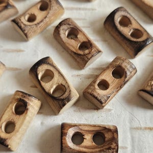 May include: A selection of rectangular wooden buttons, each with two holes. The buttons display a natural wood grain with darker, burnt edges. They are arranged on a light-coloured, textured surface.