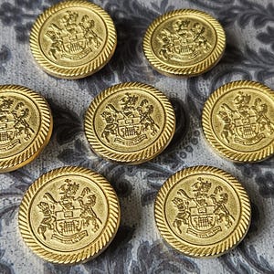 Puede incluir: Botones redondos dorados con un diseño de escudo detallado. Los botones tienen un borde en forma de cuerda y están dispuestos sobre una superficie estampada. Estos botones decorativos son adecuados para proyectos de costura.