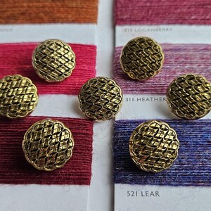 May include: Close-up of six antique gold-colored buttons with a woven design. The buttons are arranged on top of colorful spools of thread in shades of red, purple, and blue. The thread spools have labels with color names and numbers.