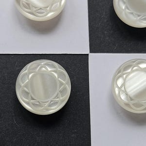 May include: Close-up of several off-white, round buttons. Each button has a floral-like design around the edge and a slightly raised centre. The buttons are arranged on a black and white chequered surface.