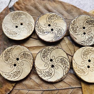 Può includere: Sei bottoni rotondi di colore chiaro con un aspetto naturale e strutturato. Ogni bottone presenta un design inciso marrone scuro con linee a spirale e forme a foglia. I bottoni hanno due fori per cucire e sono esposti su una foglia marrone.