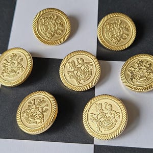 Puede incluir: Siete botones dorados con un diseño de escudo detallado y un borde en forma de cuerda. Los botones redondos están dispuestos sobre una superficie a cuadros en blanco y negro. Diseño clásico para ropa.