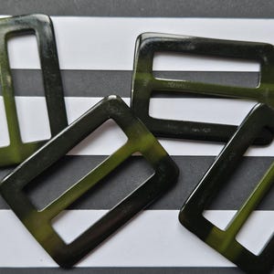 Puede incluir: Cuatro hebillas rectangulares de color verde oscuro con esquinas redondeadas dispuestas sobre un fondo a rayas blancas y negras. Las hebillas tienen un acabado brillante y una sutil variación de color, con toques de verde oliva.