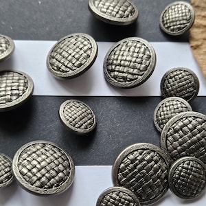 May include: A collection of silver-toned buttons with a woven texture. The buttons vary in size and have a detailed, textured surface. The buttons are arranged on a black and white striped surface.