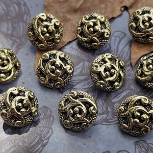May include: A collection of nine round, metallic gold-toned buttons featuring a raised, swirling filigree pattern set against a dark, textured background. Each button displays a polished, reflective surface with intricate, interconnected scrollwork.