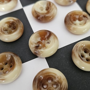 Könnte beinhalten: Eine Sammlung runder Knöpfe mit jeweils vier Löchern. Die Knöpfe haben ein marmoriertes Aussehen mit einer Mischung aus Creme- und Brauntönen. Sie sind auf einer schwarz-weiß karierten Oberfläche angeordnet.