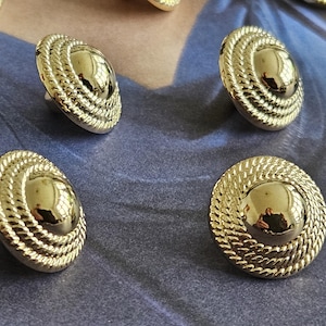 May include: A collection of gold-coloured buttons. Each button has a shiny, domed centre and a rope-like textured border. The buttons are oval and appear to be metal. They are arranged on a blue surface.