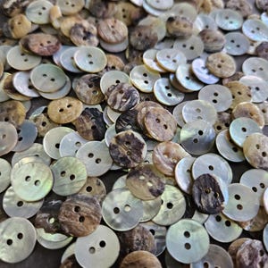 May include: A collection of round buttons with two holes each. The buttons are made of a pearlescent material, with colors ranging from white and light blue to brown and black. The buttons are scattered across a dark surface.