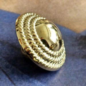 May include: A gold button with a rope-style edge and a smooth, domed centre. The button is set against a blue background, showing its shiny, reflective surface. This decorative button is suitable for various sewing projects.