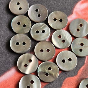 May include: A collection of round, iridescent buttons with two holes each. The buttons are a silvery-gray color and appear to be made of a pearlescent material. The buttons are scattered on a patterned background.