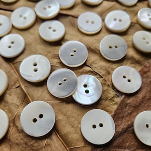 May include: A collection of round, off-white buttons with two holes each, scattered on a brown, textured surface. The buttons have a pearlescent sheen, reflecting light. Ideal for sewing and craft projects.